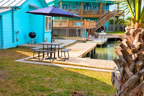 Canal-front backyard in Rockport, Texas with wooden dock, picnic table and umbrella, blue coastal home, and water access for boating and fishing.