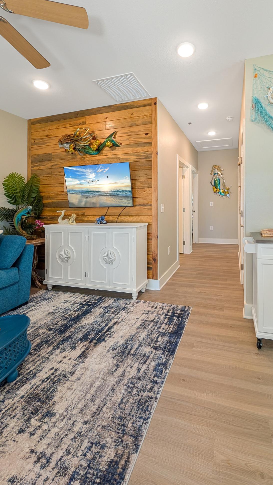 Living area with a wood accent wall, coastal decorations, TV on a cabinet, blue sofa, and hallway leading to other rooms with wood-look flooring.