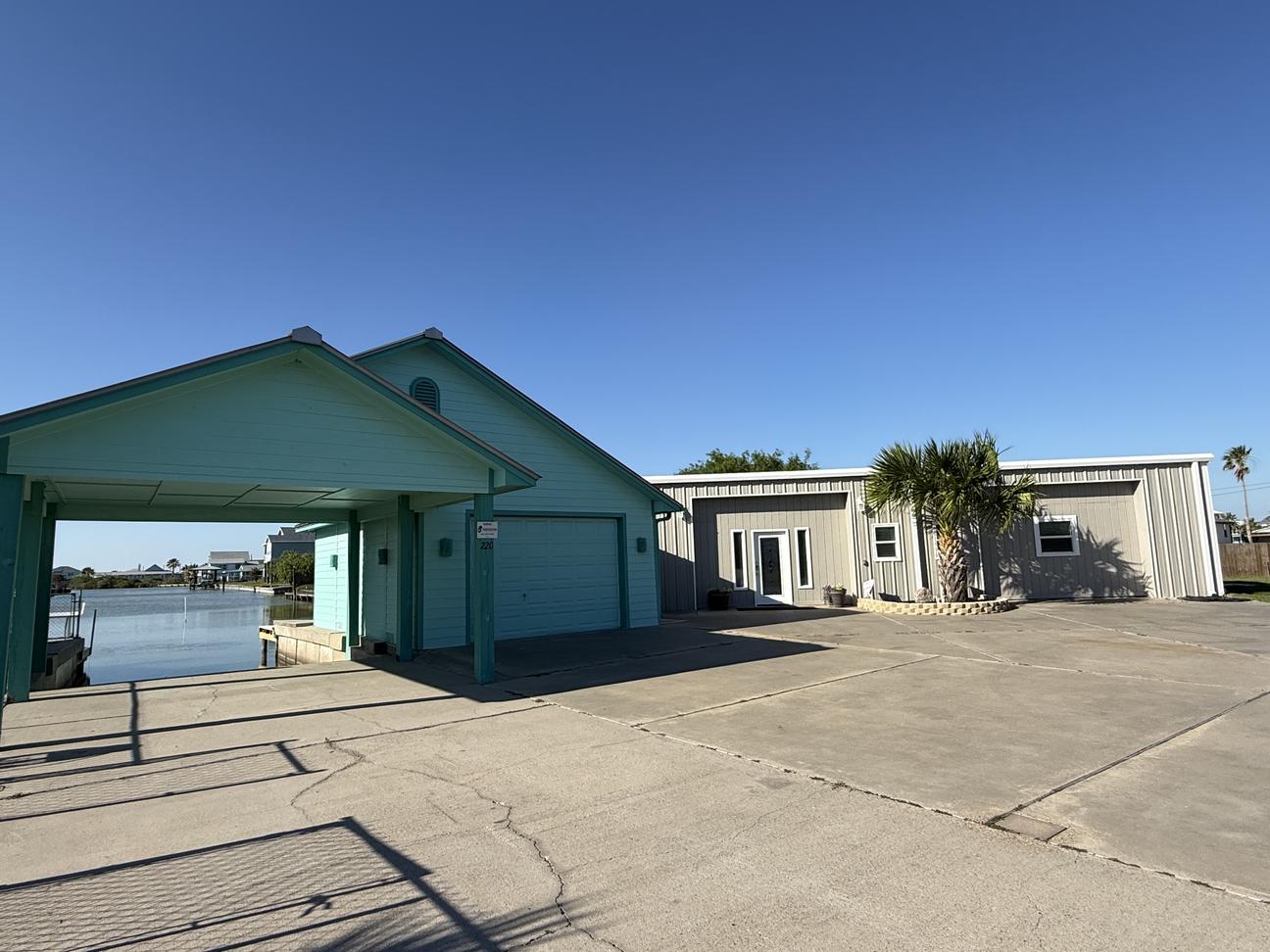 Waterfront coastal property with covered parking, garage space, large driveway, and canal access beside a modern beach-style home under a clear blue sky.