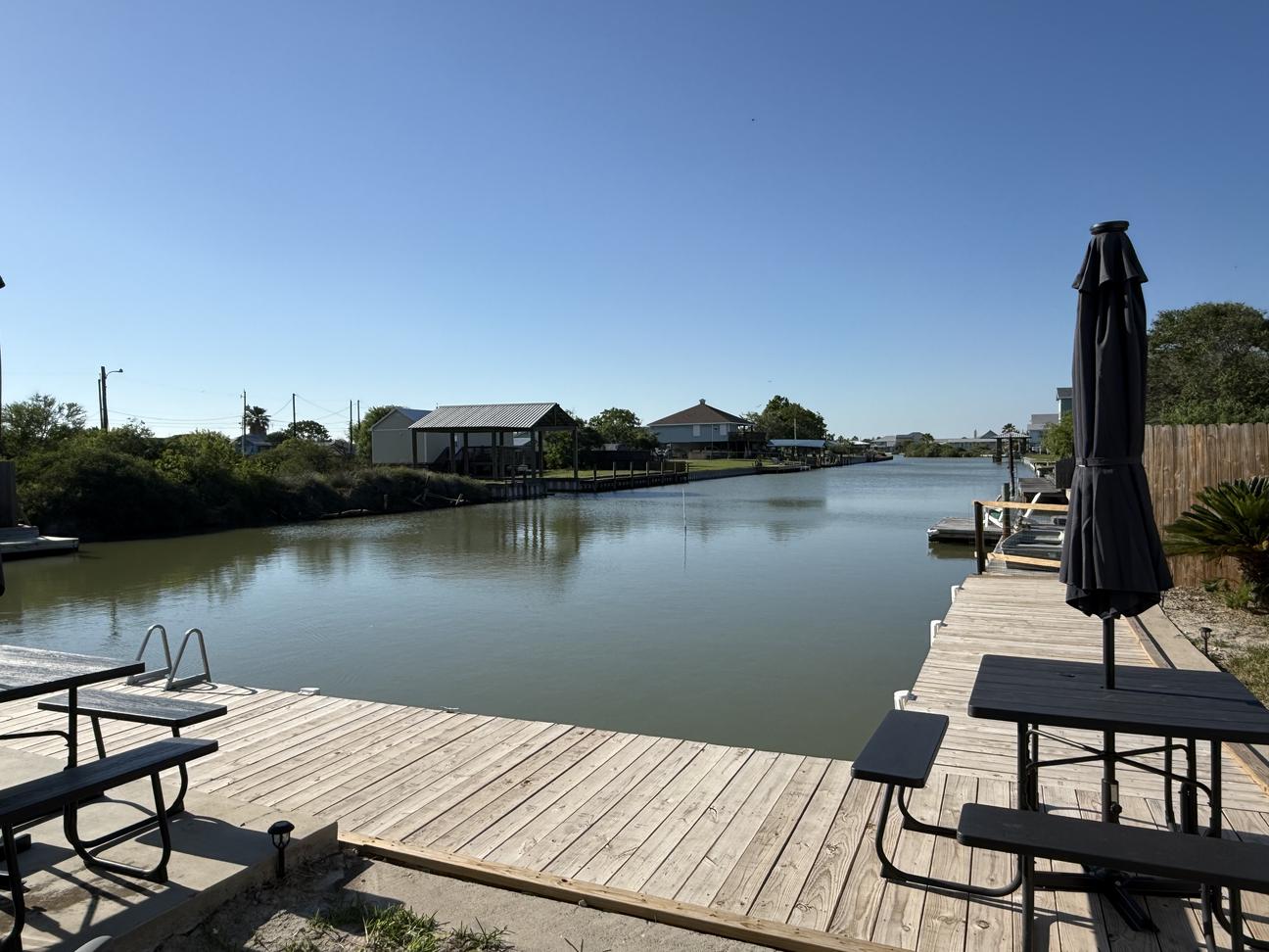 Private waterfront dock on a quiet canal with outdoor picnic tables, umbrella seating, and calm water views surrounded by coastal homes under a clear blue sky.