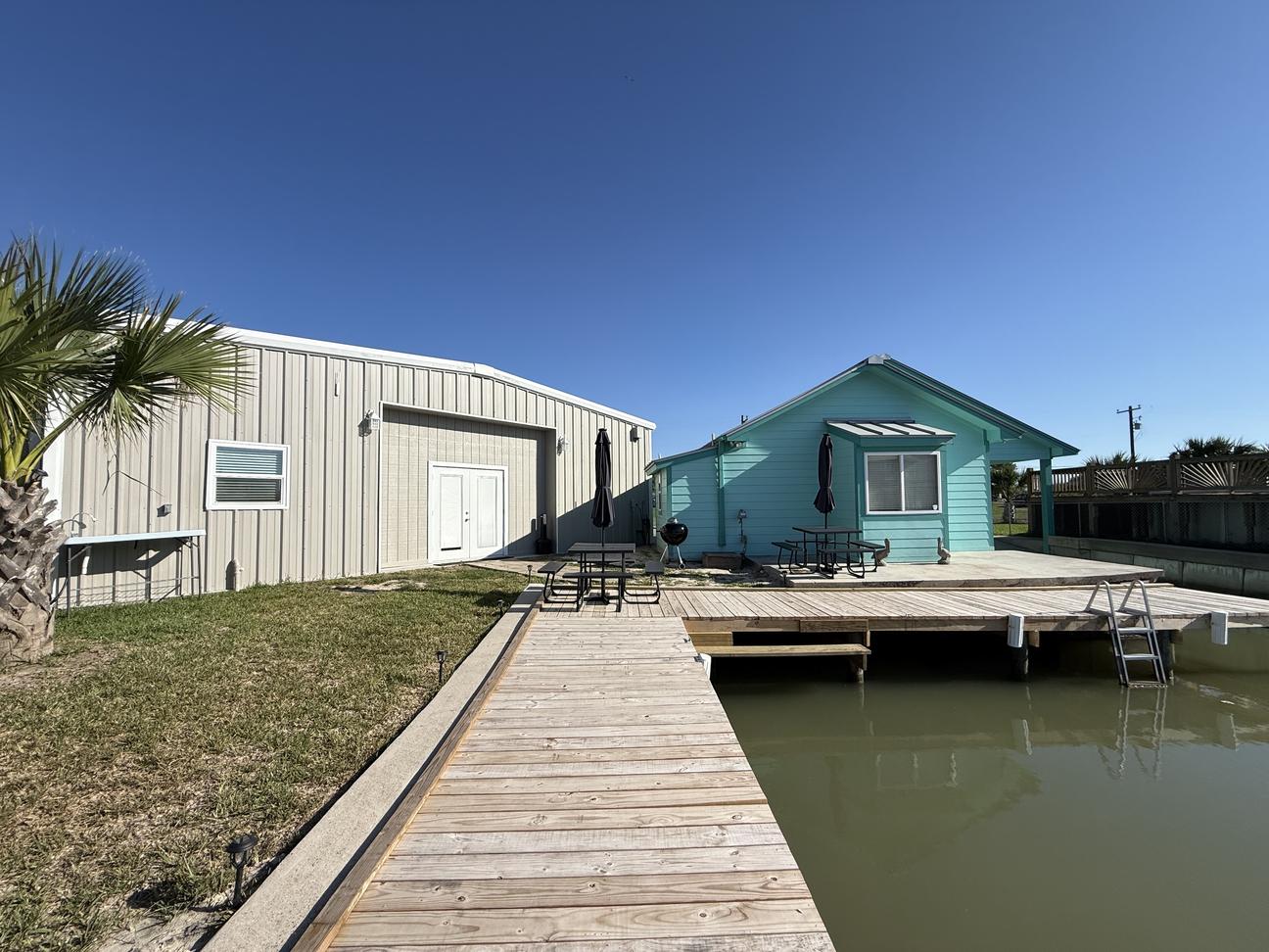 Canal-front coastal property with private dock, waterfront deck seating, outdoor dining area, and direct water access beside a bright turquoise beach-style home under a clear blue sky.