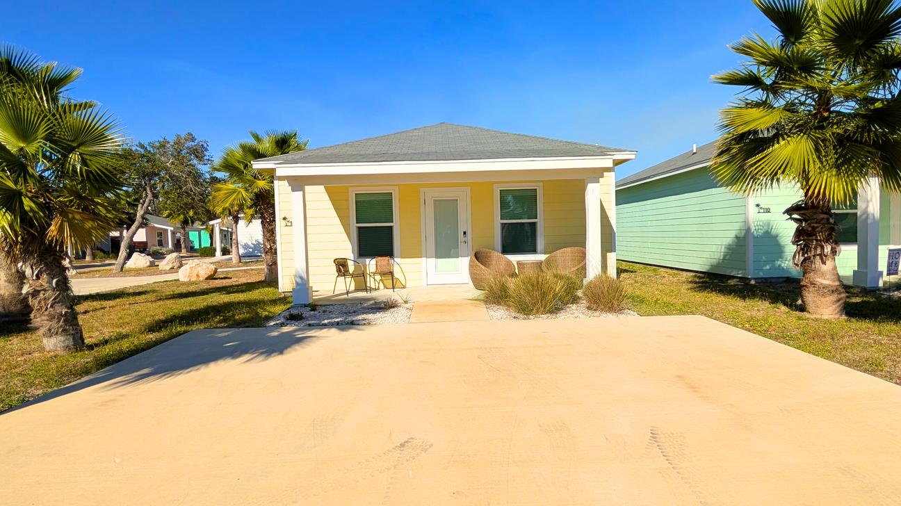 Single-story yellow coastal cottage with a small front porch, chairs, palm trees, and a wide driveway under a clear blue sky.
