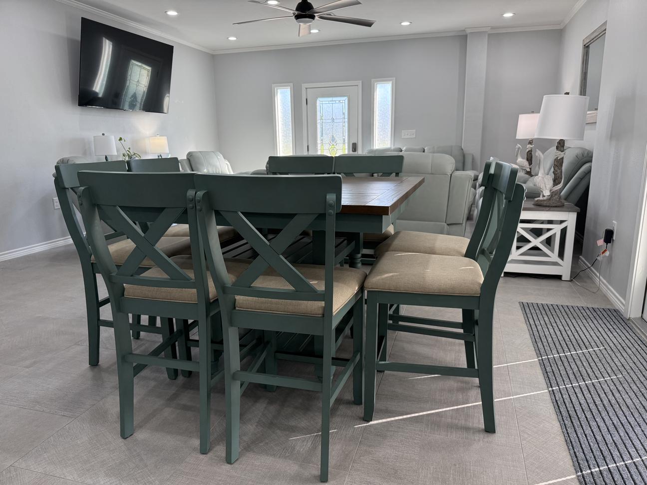 Coastal-style dining and living area with seating for six, soft neutral décor, comfortable recliners, large wall-mounted TV, and bright open-concept layout in a modern vacation rental.