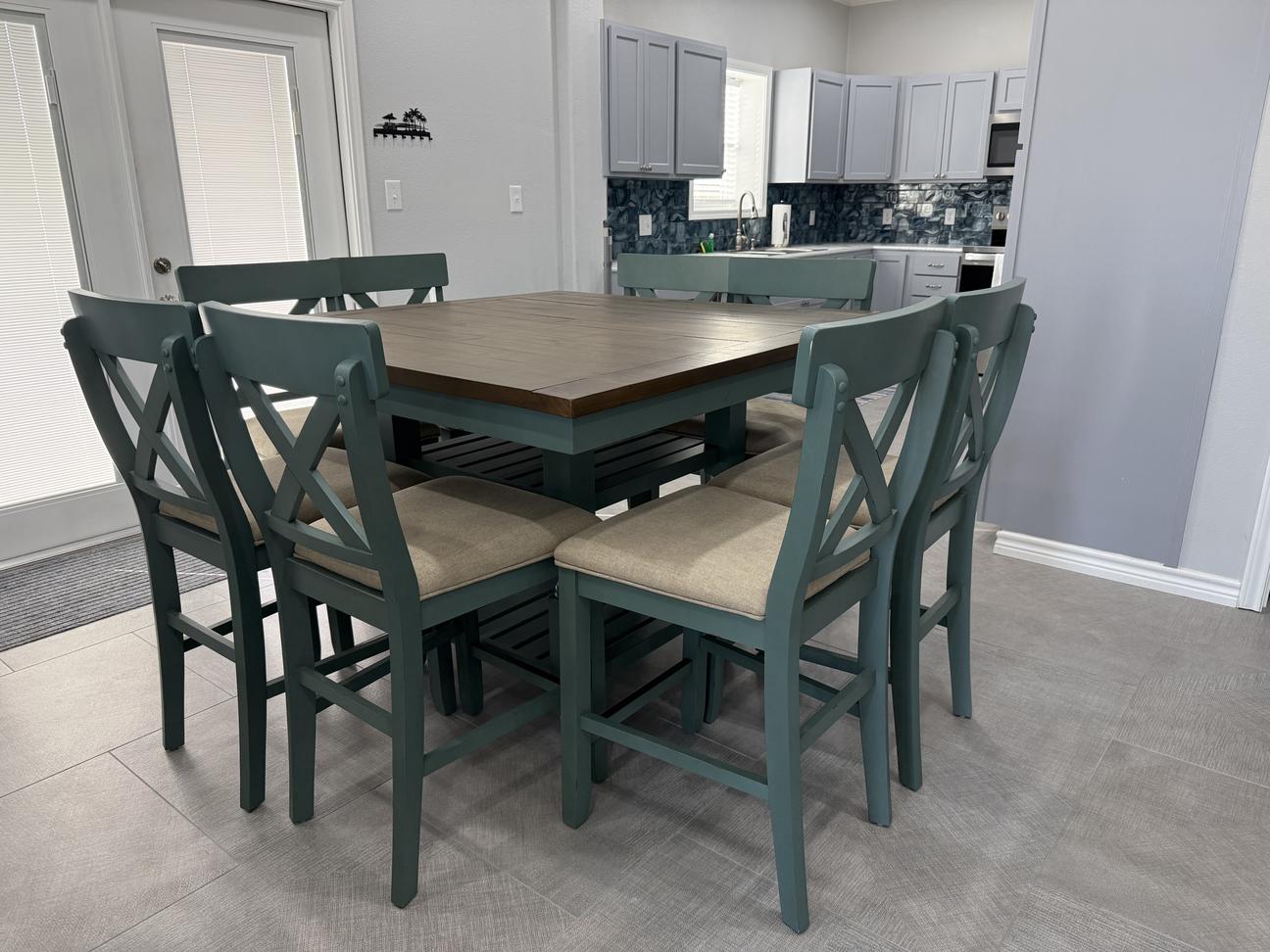 Coastal-style dining area with seating for six, blue-green chairs, wood dining table, and open view into a modern kitchen with beach-inspired finishes in a bright vacation rental.