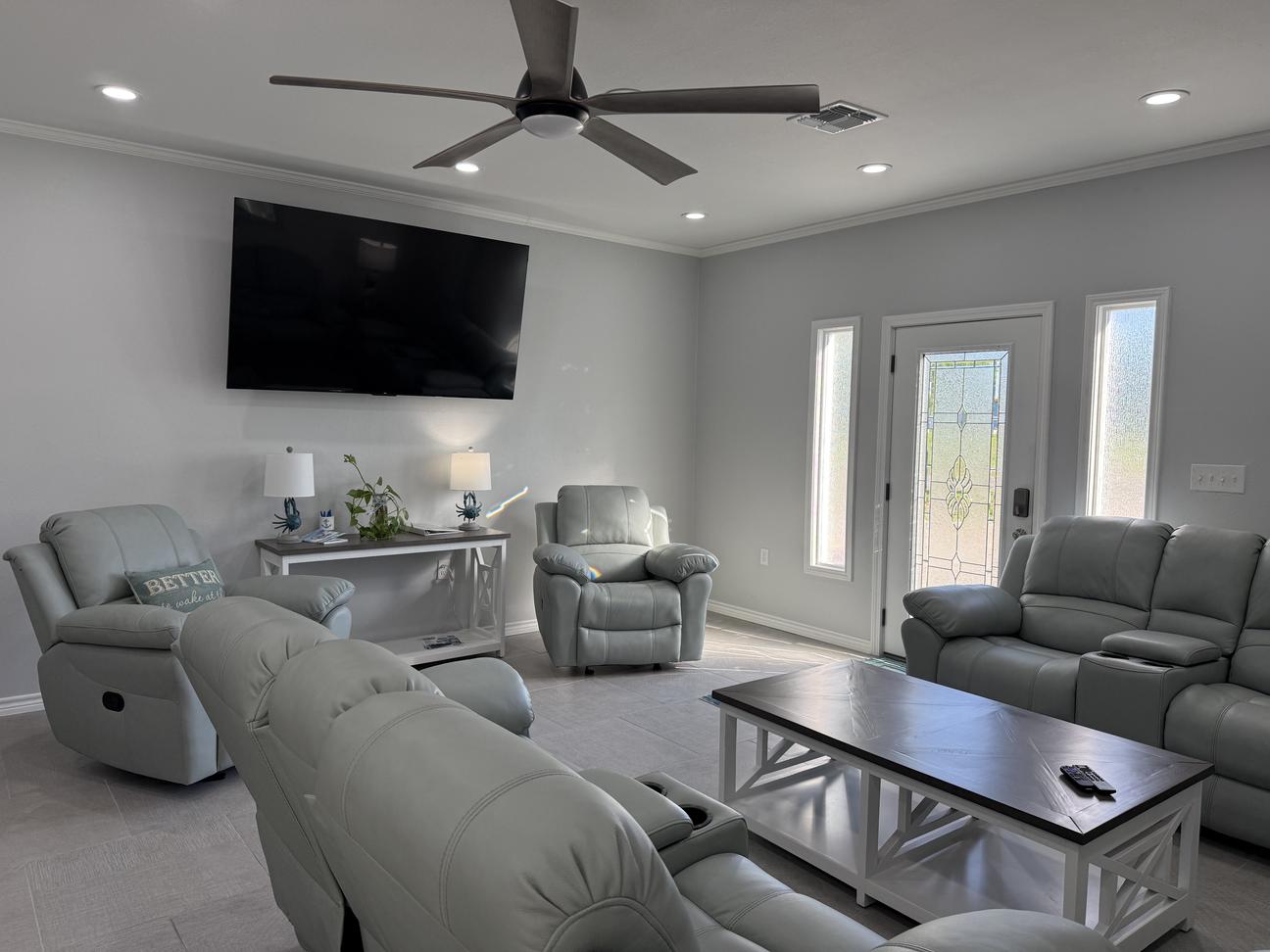 Spacious coastal-style living room with plush reclining seating, large wall-mounted TV, ceiling fan, bright natural light, and soft neutral décor in a modern vacation rental.