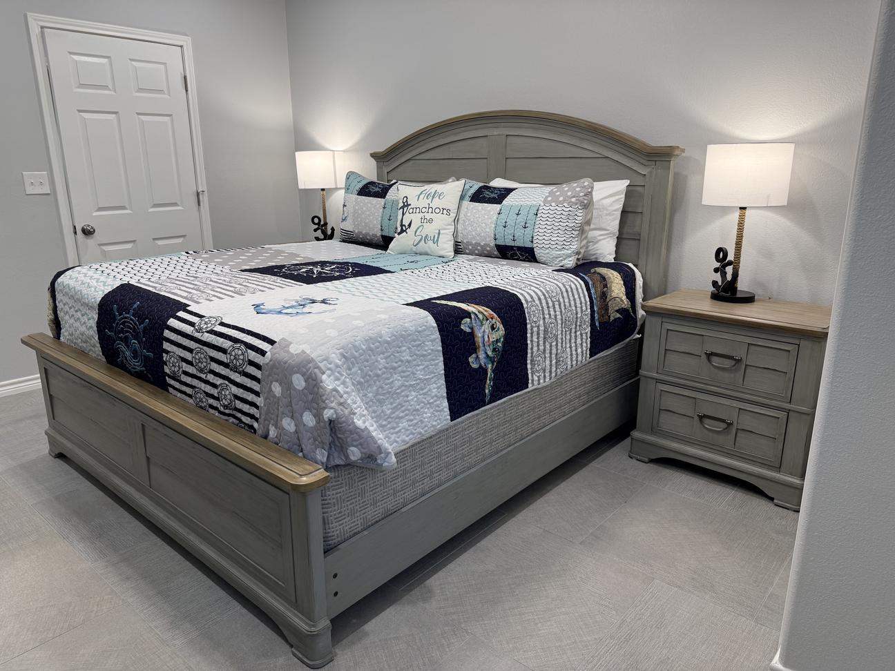 Spacious coastal-style bedroom featuring a king bed with nautical-themed bedding, matching gray wood furniture, bedside lamps with anchor accents, and light neutral décor.