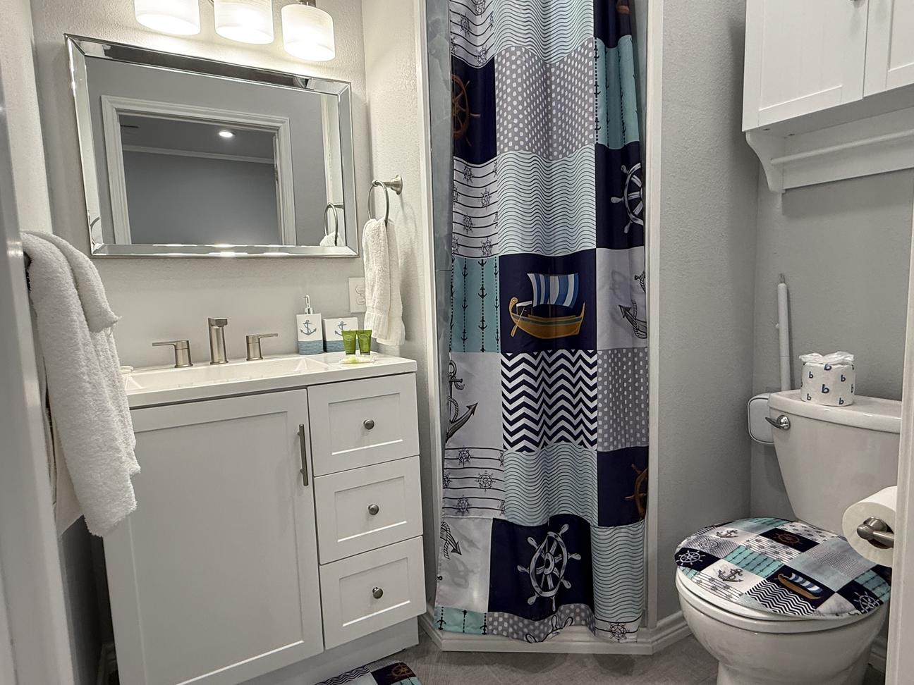 Bright coastal-themed bathroom with white vanity, nautical shower curtain featuring anchors and ships, modern fixtures, soft lighting, and beach-inspired décor accents.