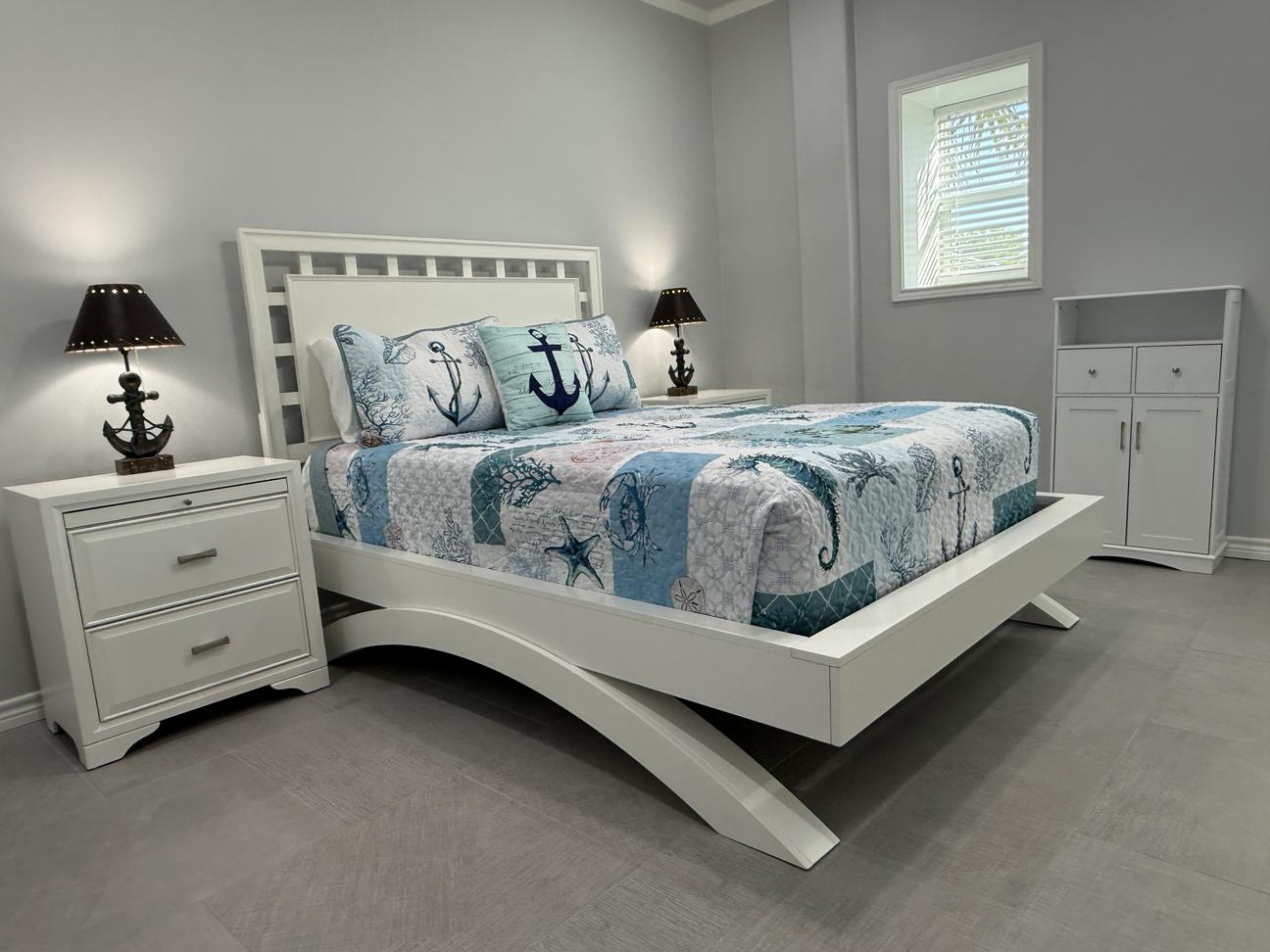 Coastal-inspired bedroom with a white bed frame, blue nautical bedding, anchor-themed accent pillows, matching nightstands with anchor lamps, and soft gray walls.