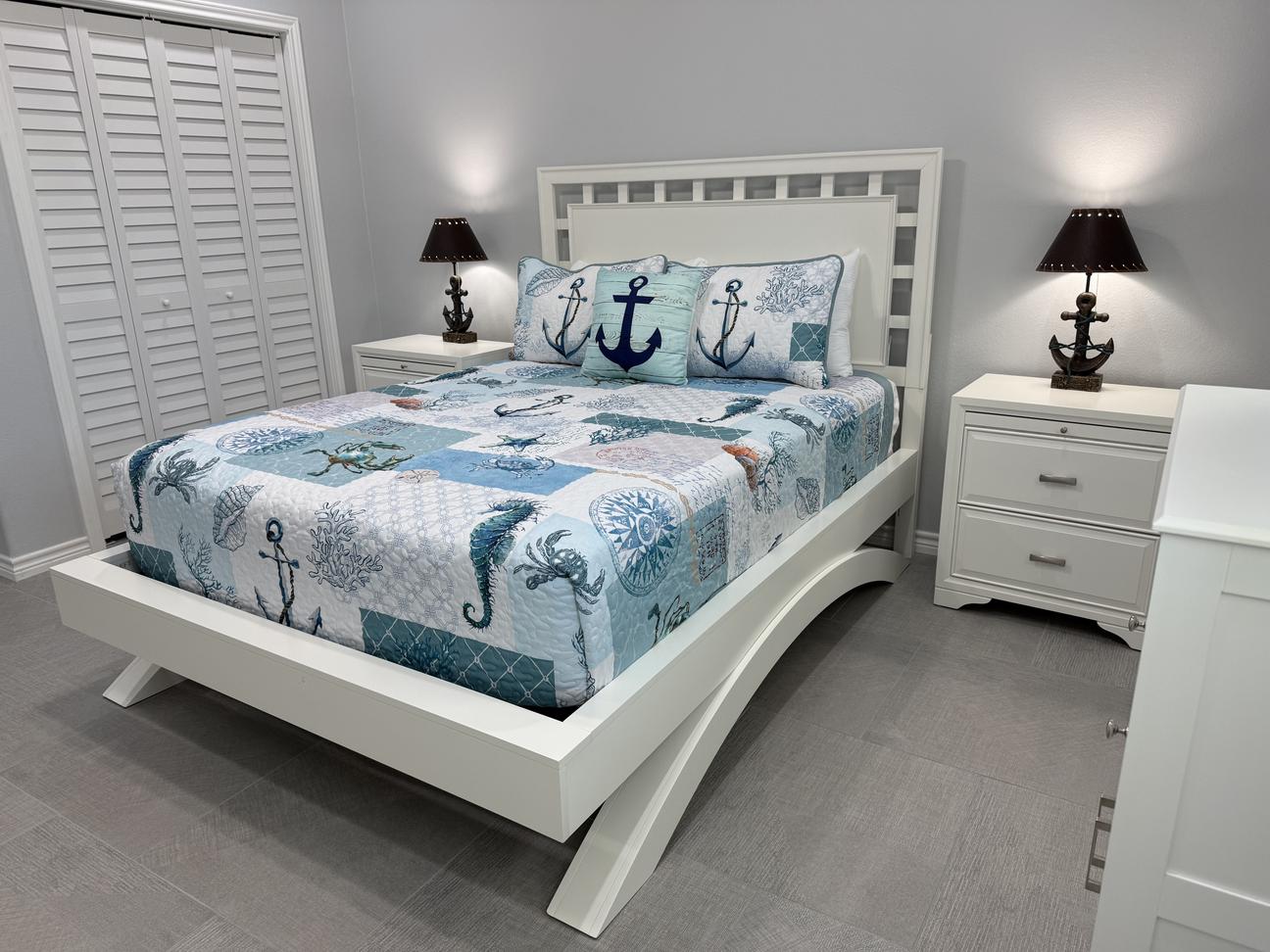 Bright coastal-themed bedroom with a white modern bed frame, nautical bedding in blue and white tones, anchor accent pillows, matching nightstands with anchor lamps, and gray tile flooring.