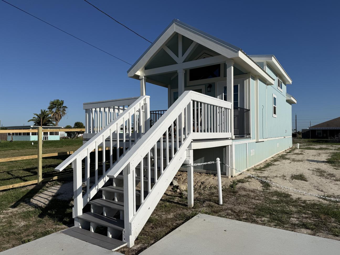 Blue tiny home with a large porch with steps and a white railing, with a blue sky and concrete parking area.