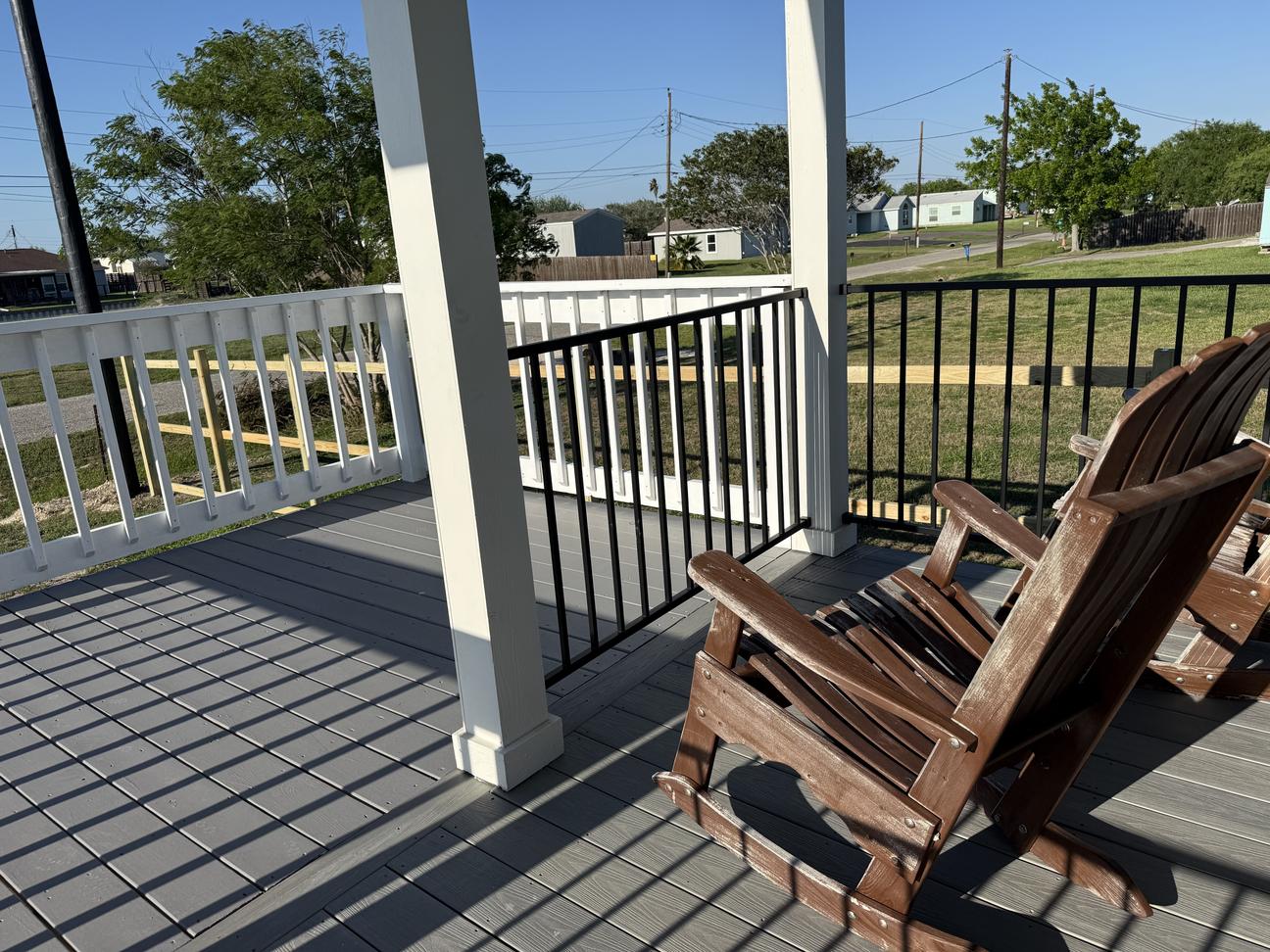 Covered outdoor deck with gray flooring, black-and-white railing, and wooden Adirondack chairs overlooking a quiet neighborhood under a clear blue sky.