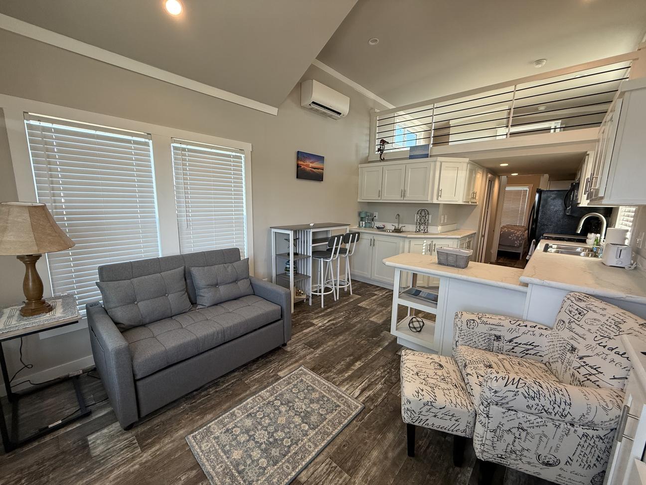 Open-concept coastal-style vacation rental interior with vaulted ceilings, loft sleeping area, white kitchen cabinetry, modern furnishings, gray loveseat, patterned accent chair, wood-look flooring, and abundant natural light throughout the space