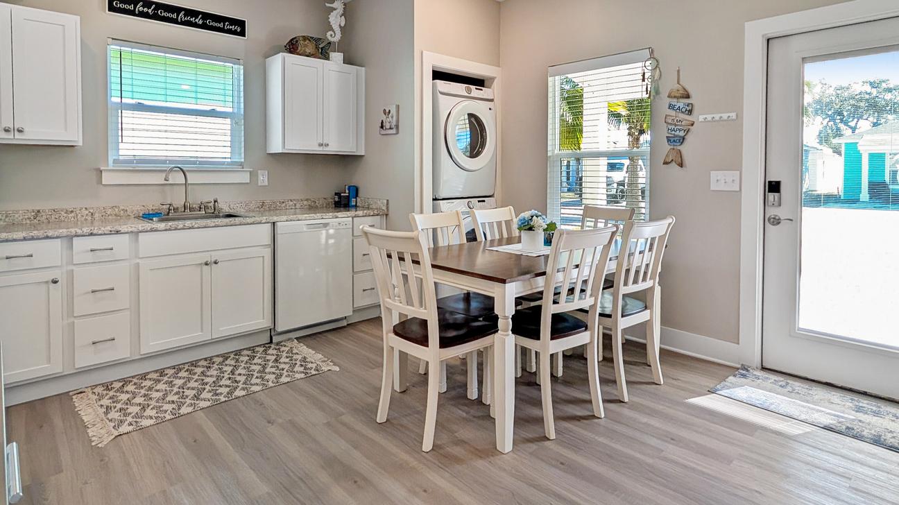 White wood dining table with seating for six, stack washer and dryer and clean kitchen with white cupboards.