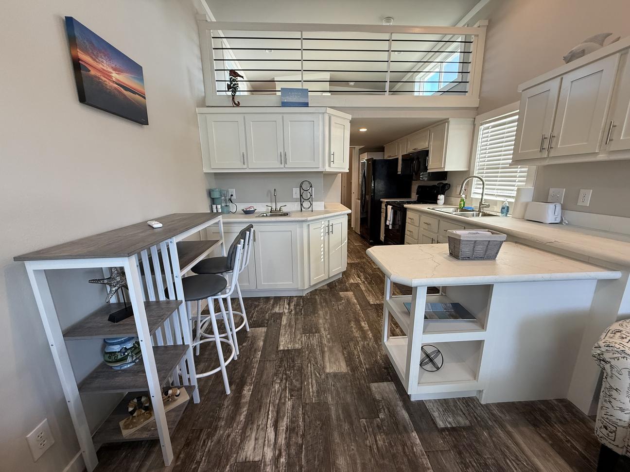 Bright coastal-style vacation rental featuring vaulted ceilings, loft sleeping area with railing, white cabinetry, breakfast bar seating for two, modern kitchen with island, and wood-look flooring in an open-concept layout.