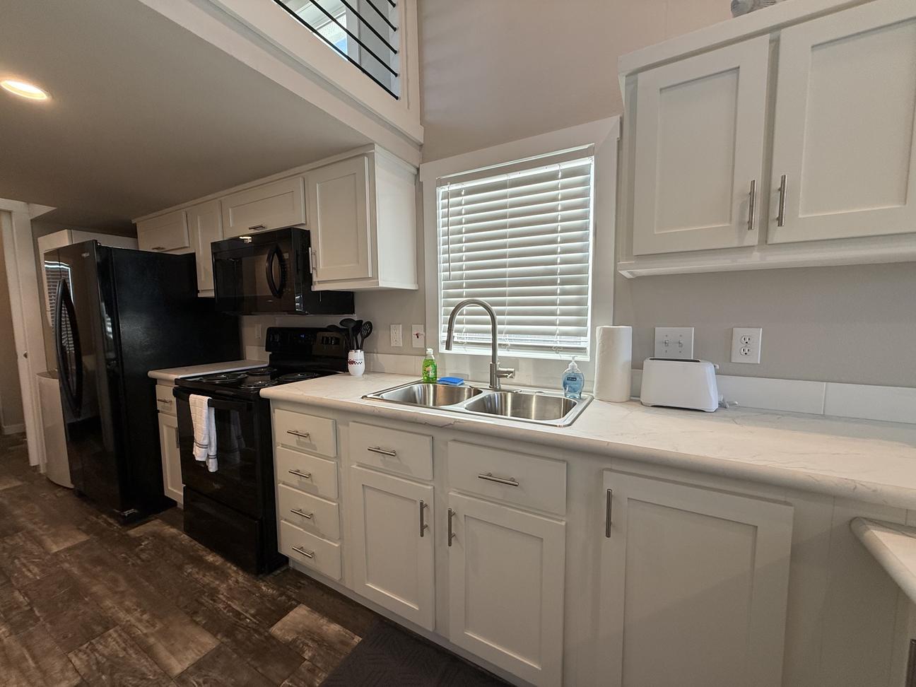 Bright vacation rental kitchen featuring white shaker cabinets, black appliances, double stainless sink, high-arc faucet, wood-look flooring, and tall ceilings with loft overlook in a clean coastal-style interior.