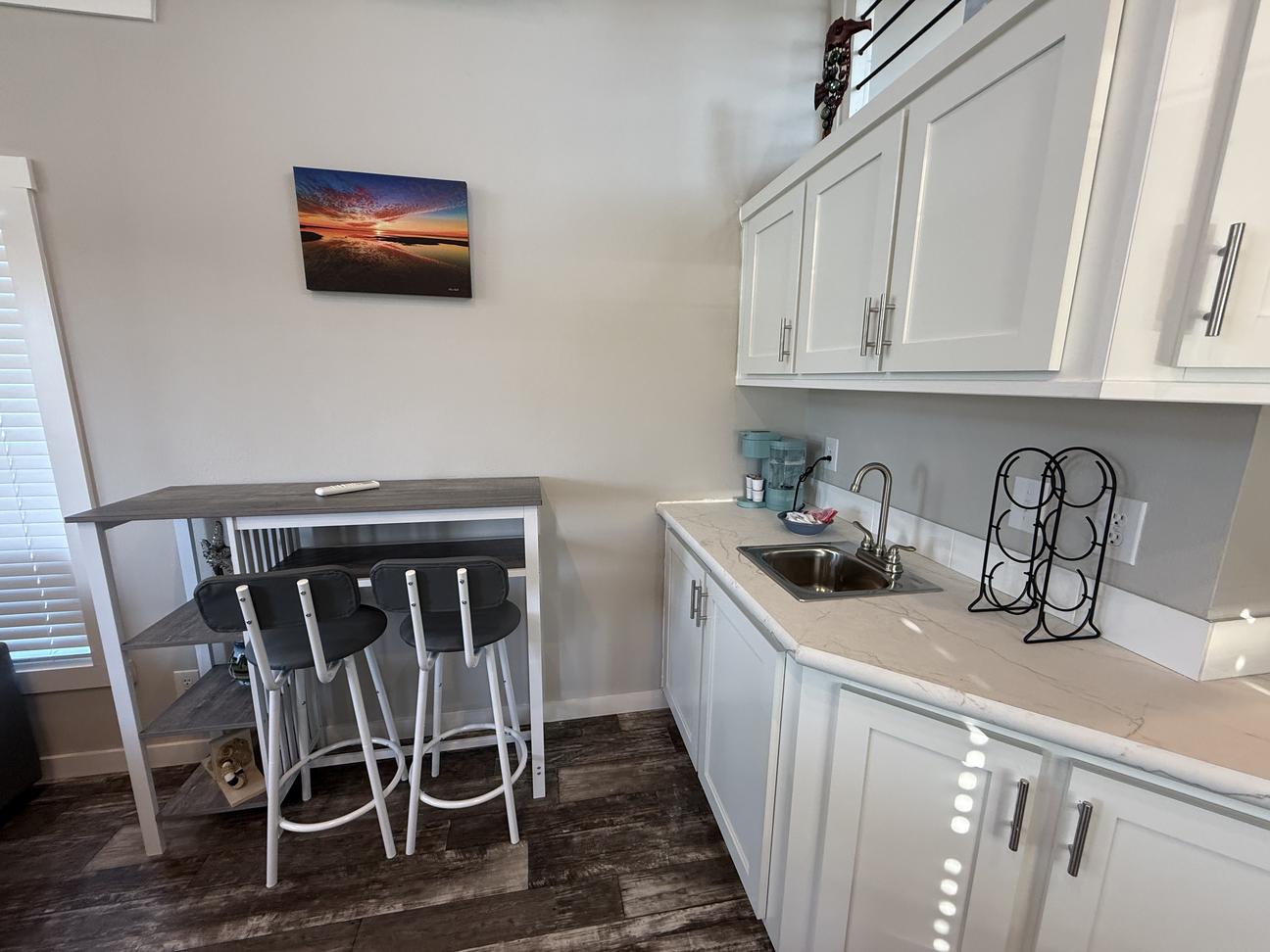 Coastal-style kitchenette area with white cabinets, small sink, breakfast bar seating for two, wood-look flooring, and beach sunset wall art in a bright vacation rental interior.