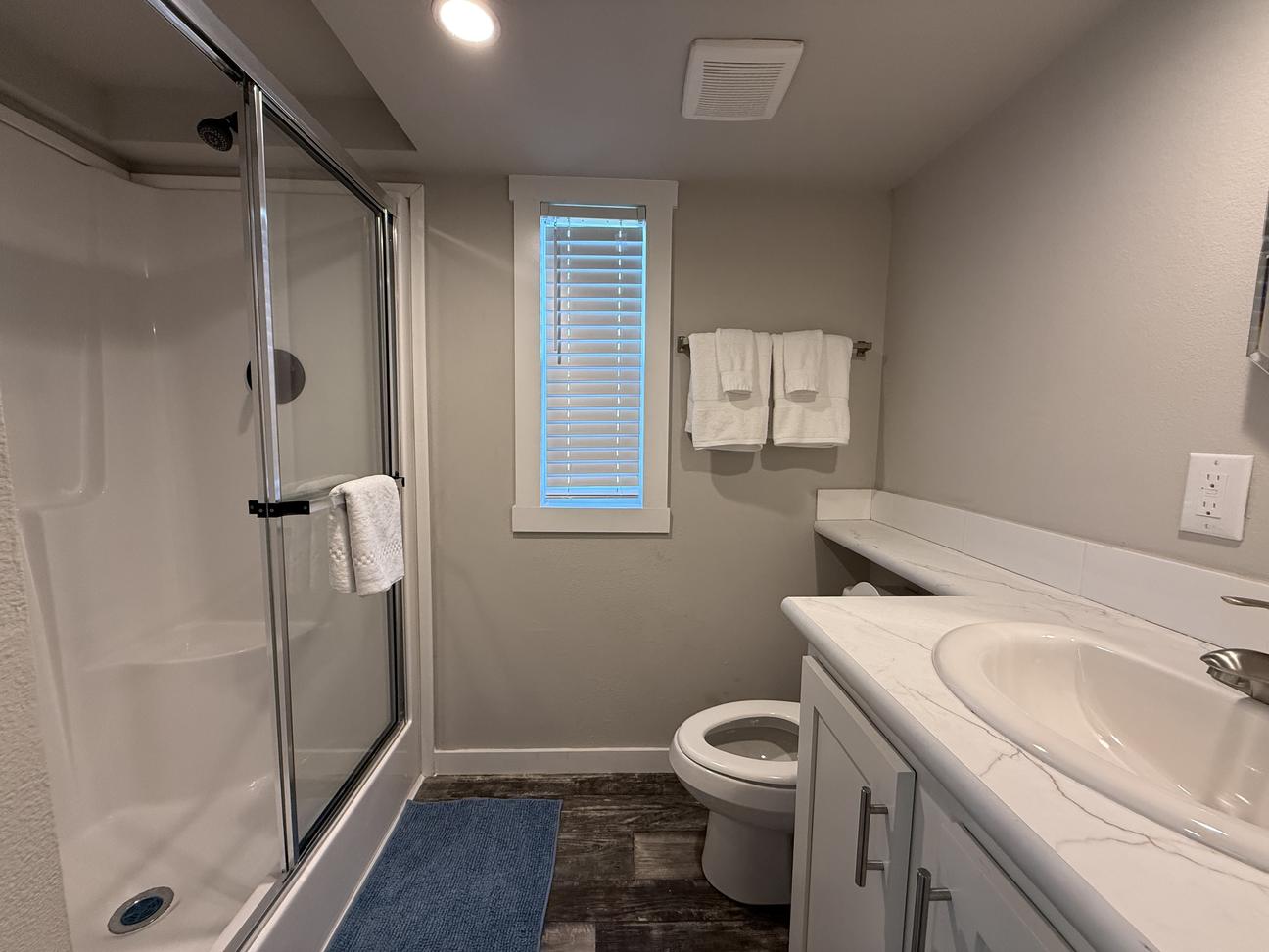 Modern vacation rental bathroom featuring a large walk-in shower with glass doors, white vanity, neutral coastal décor, wood-look flooring, and fresh white towels.