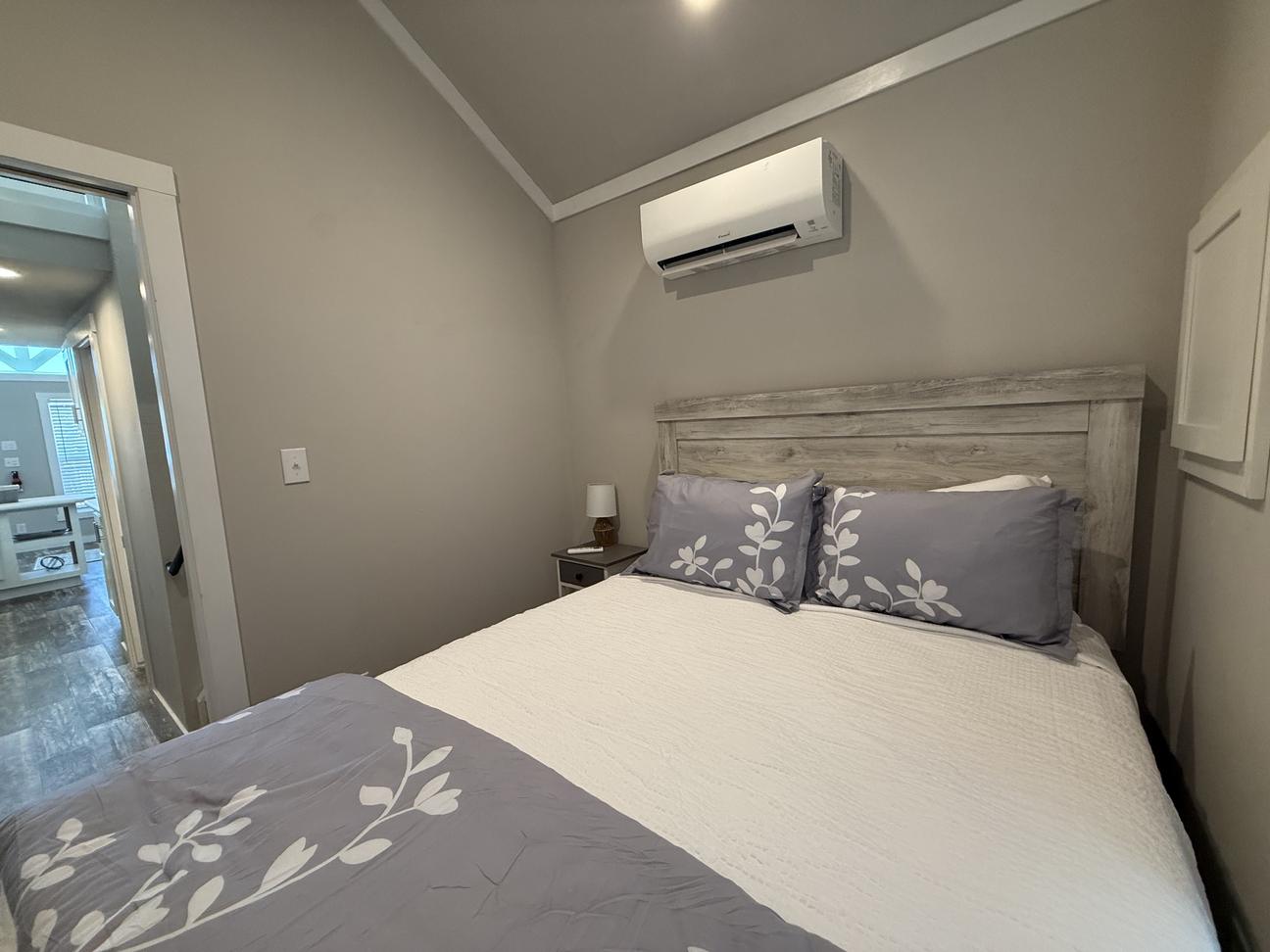 Bright coastal-style bedroom featuring a queen bed with gray and white bedding, wood-look furniture, bedside table, vaulted ceiling, window with blinds, and warm neutral décor.