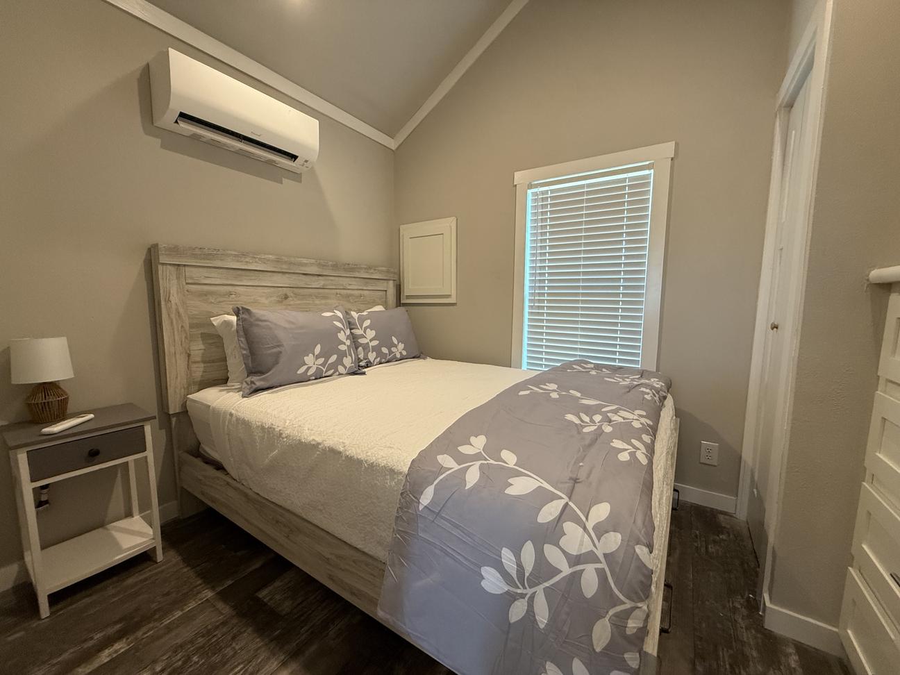 Bright coastal-style bedroom featuring a queen bed with gray and white bedding, wood-look furniture, bedside table, vaulted ceiling, window with blinds, and warm neutral décor.