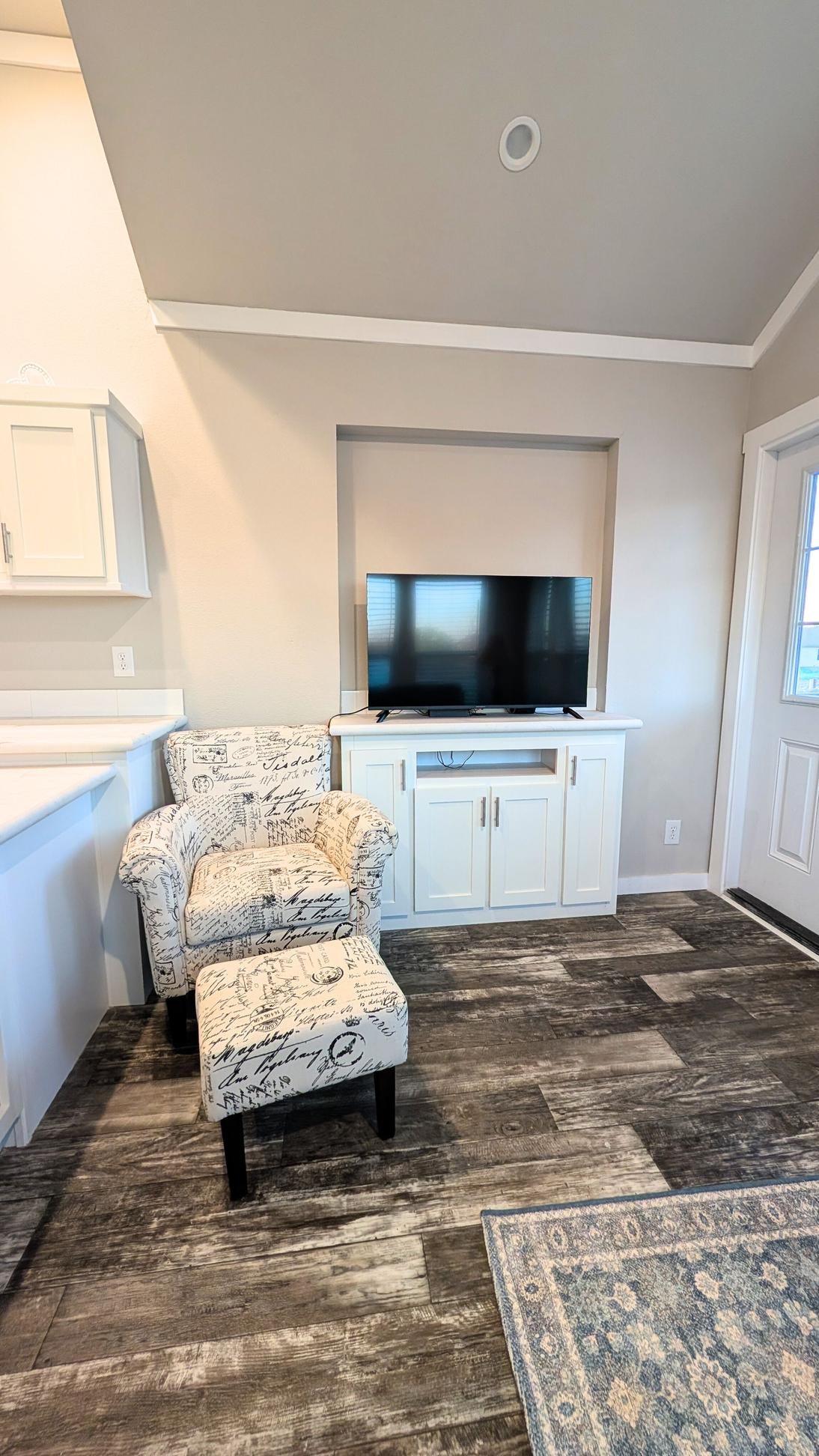 Small living area with a patterned armchair and ottoman, a flat-screen TV on a built-in cabinet, light neutral walls, and wood-style flooring.