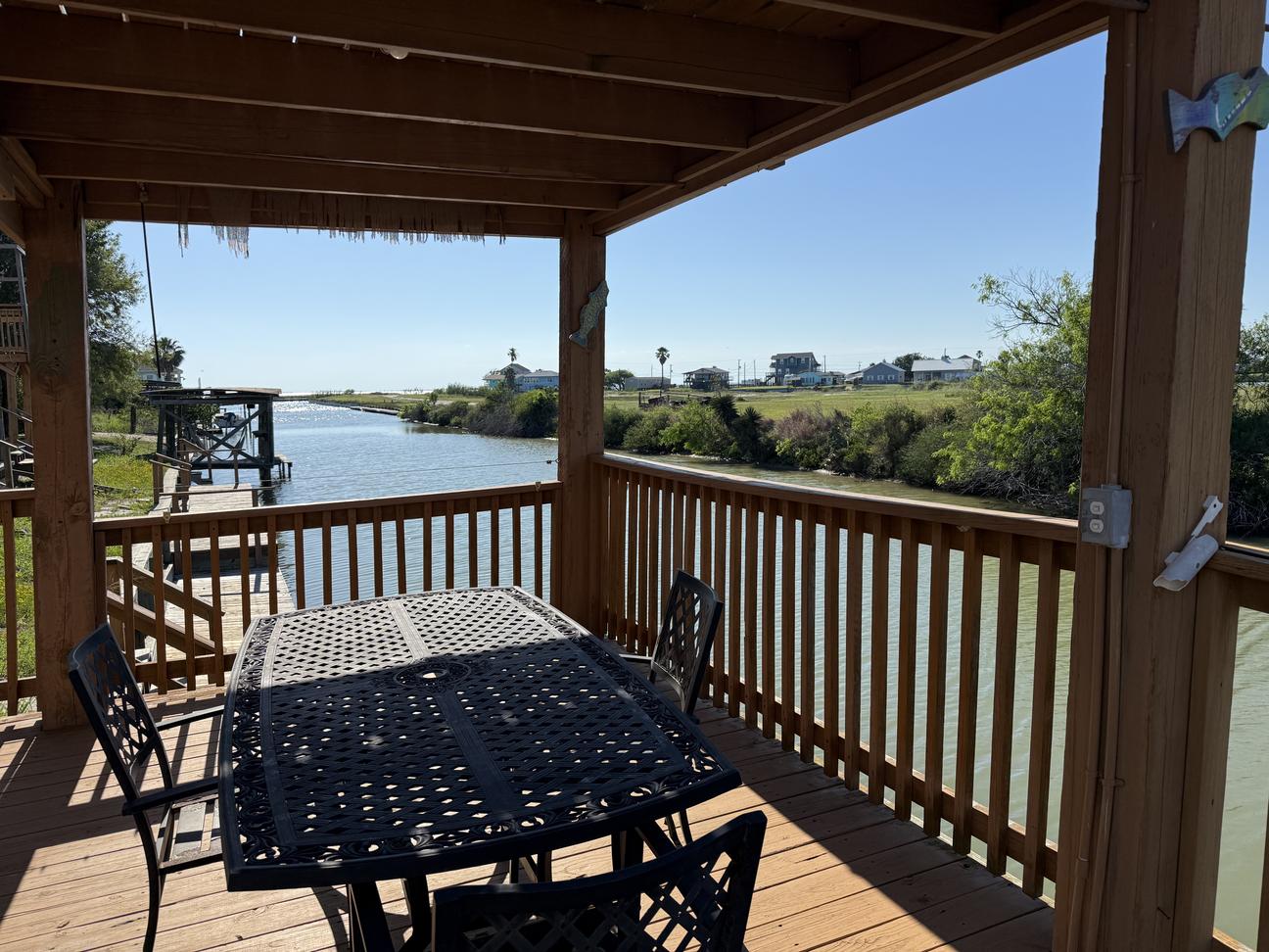 Covered waterfront deck overlooking a calm canal with outdoor dining table, wooden railing, and views of nearby coastal homes and open water under a clear blue sky.
