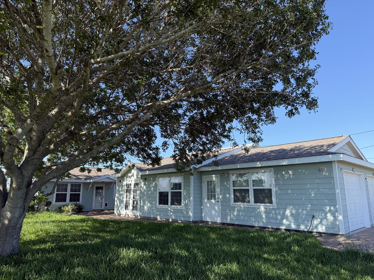 Single-story coastal home with light blue siding, large front yard, mature shade tree, and covered entryway on a sunny day.
