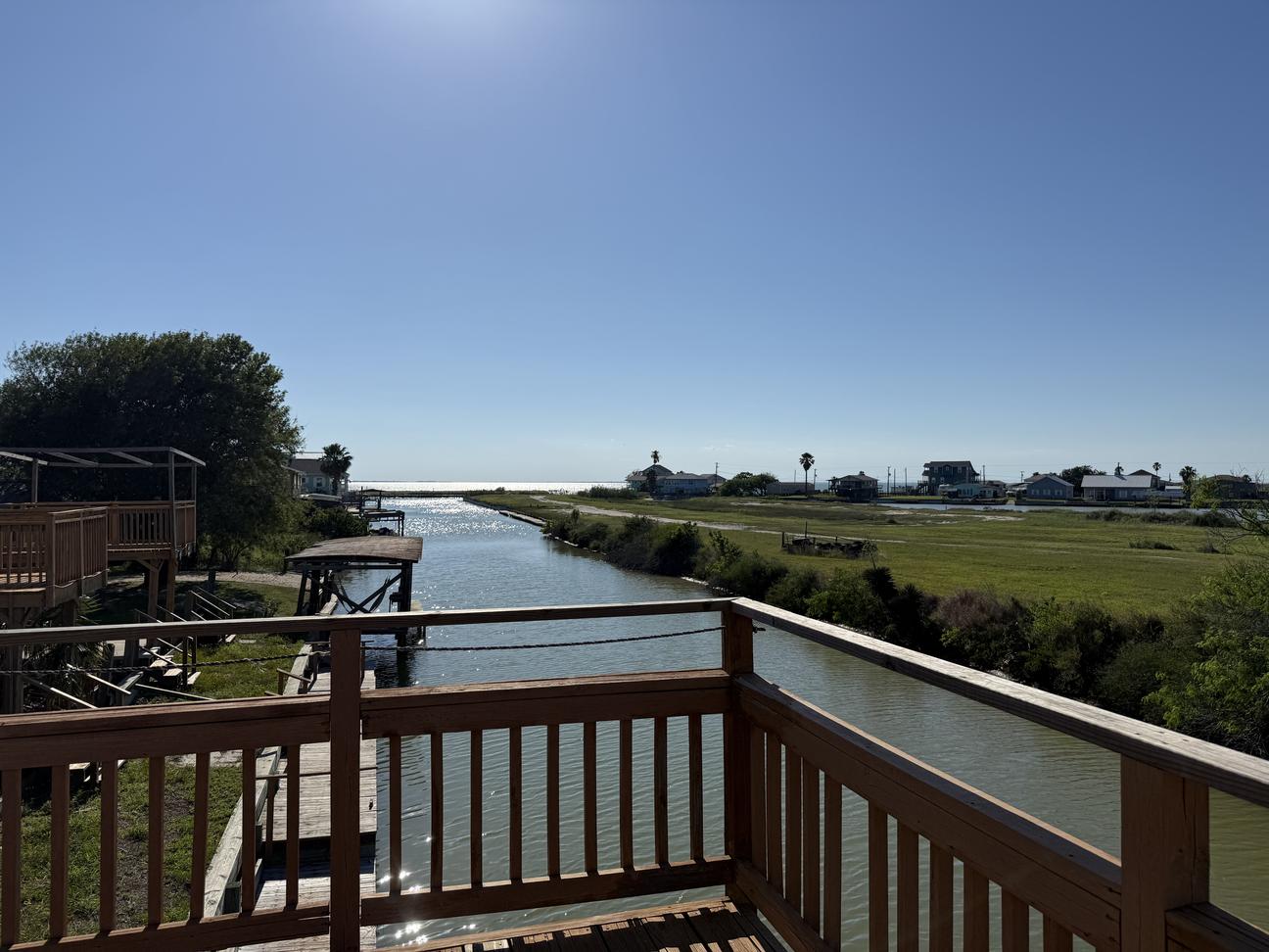 View from a waterfront deck overlooking a canal leading toward open water, with neighboring docks, grassy shoreline, and bright blue sky on a sunny coastal day.