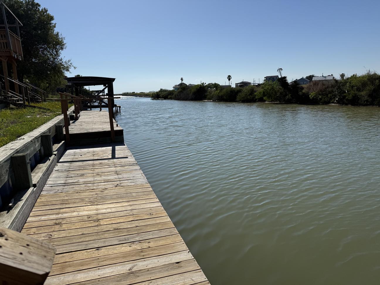 Long wooden dock along a quiet canal with calm water, nearby boat lift, and coastal homes in the distance beneath a clear sunny sky.