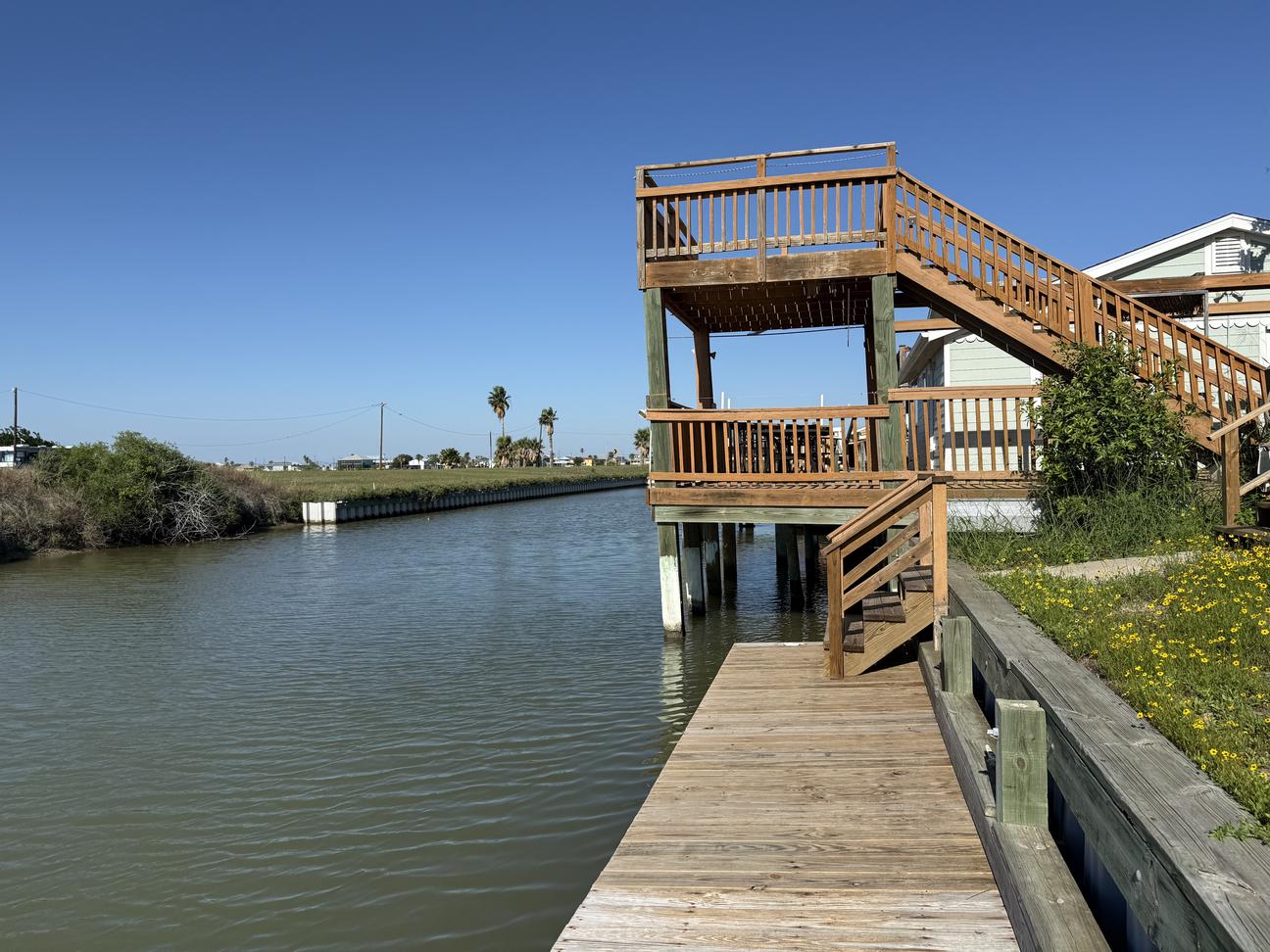 Wooden multi-level deck overlooking the water at sunset, with a table and chairs, railing, and stairs leading to an upper viewing platform.