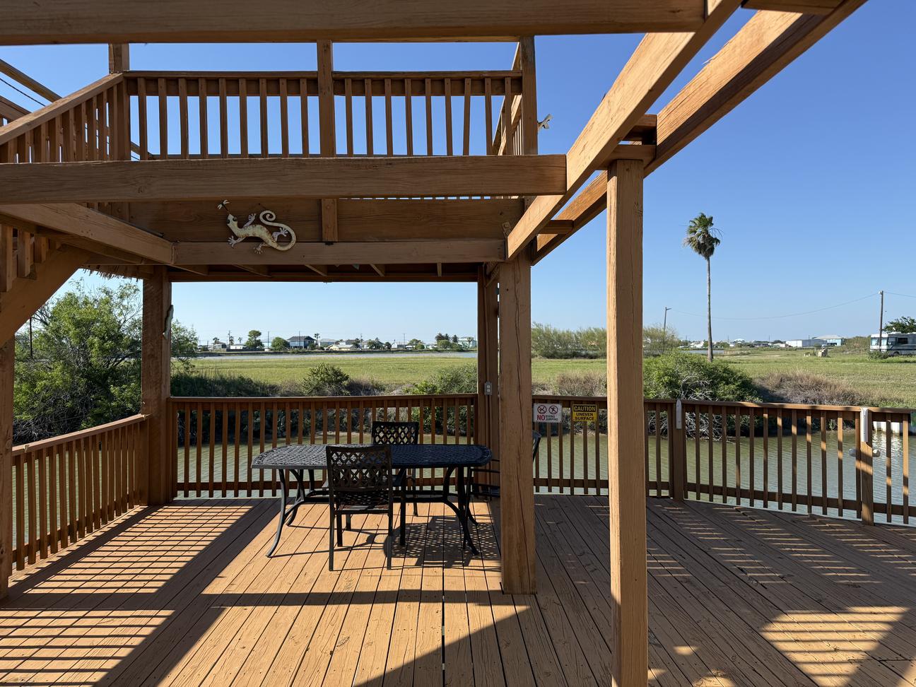 Spacious multi-level waterfront deck with wood railings, outdoor dining table, canal views, and open coastal landscape under a bright blue sky.