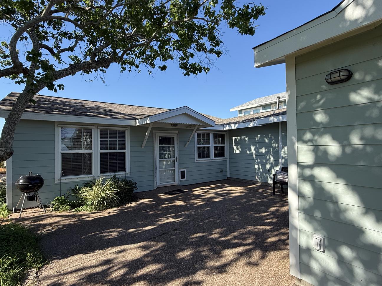 Single-story coastal home with light blue siding, large front yard, mature shade tree, and covered entryway on a sunny day.