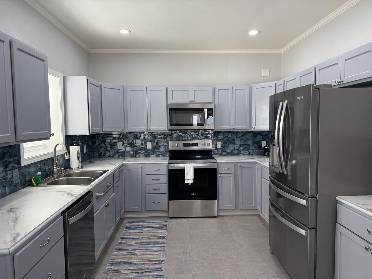 Modern coastal kitchen with gray cabinetry, stainless steel appliances, blue ocean-inspired backsplash, white countertops, and spacious U-shaped layout.