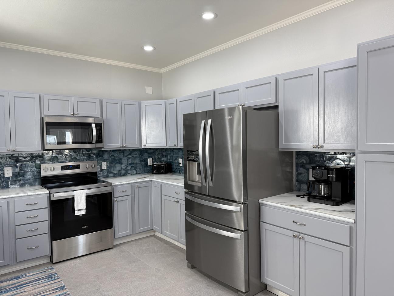 Spacious modern kitchen with light gray cabinets, stainless steel appliances, blue coastal-style backsplash, white countertops, and bright recessed lighting.