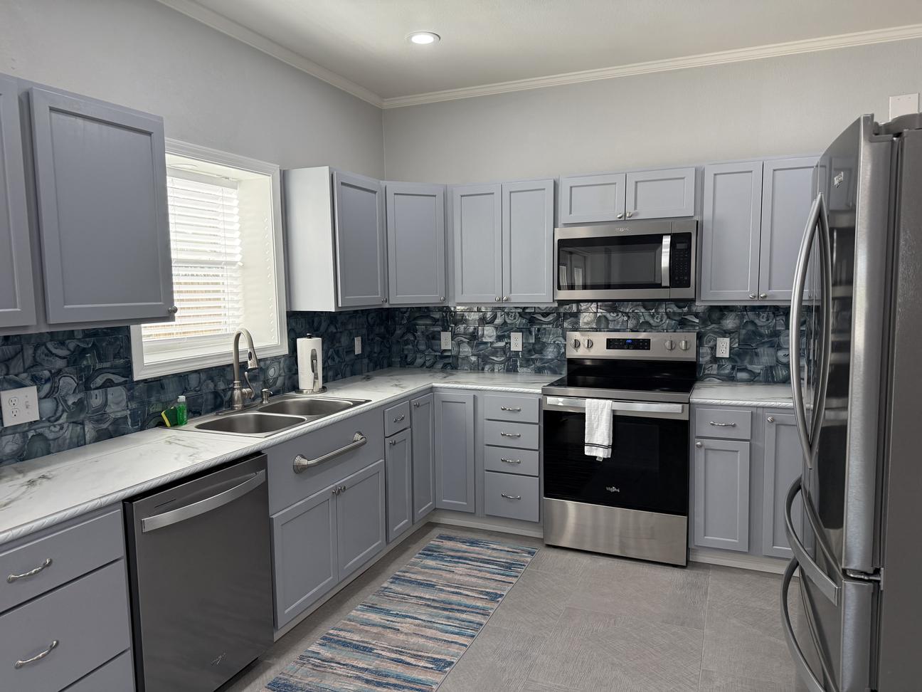 Modern coastal kitchen with gray cabinetry, stainless steel appliances, blue patterned backsplash, white countertops, double sink, and bright natural lighting.