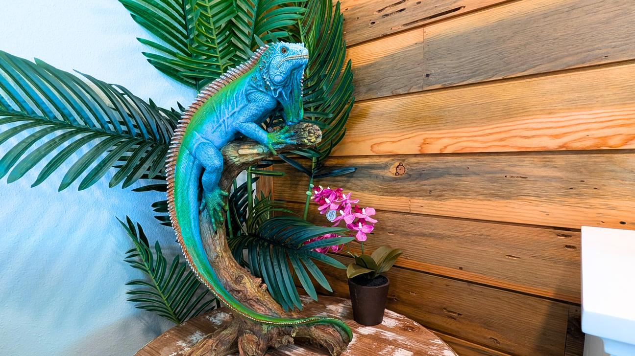 Decorative blue iguana statue on a wooden base surrounded by tropical leaves and a small pink flower on a table against a wood accent wall.