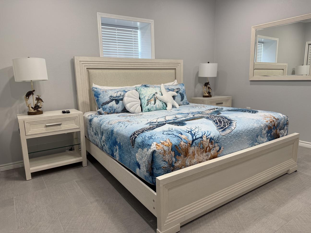 Coastal-inspired bedroom with a light wood bed frame, sea turtle and ocean-themed bedding, decorative beach pillows, matching nightstands with lamps, and soft gray walls.
