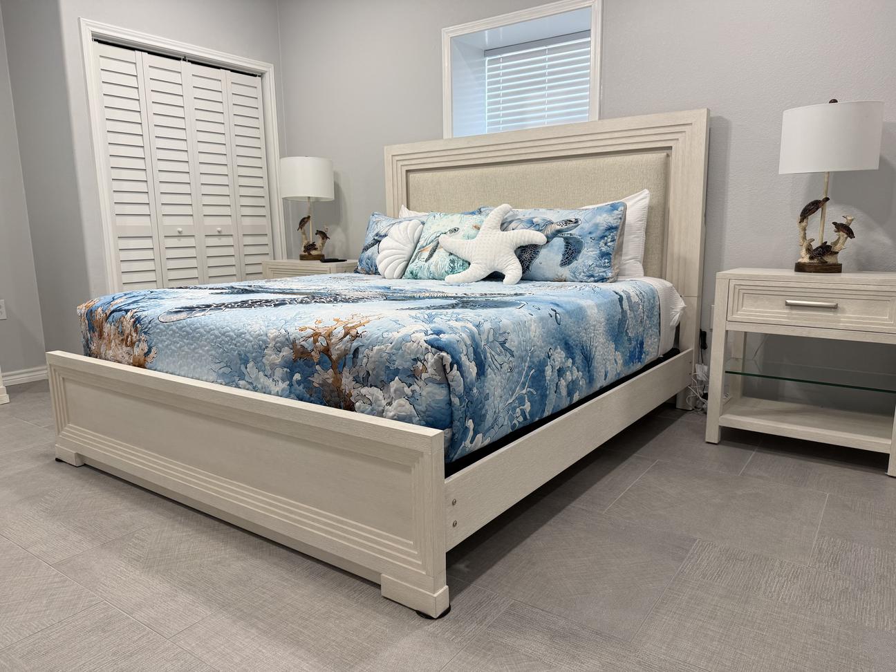 Coastal-style bedroom with a light wood bed frame, blue ocean-themed bedding, decorative starfish pillow, matching white nightstands with lamps, and light gray walls with tiled flooring.