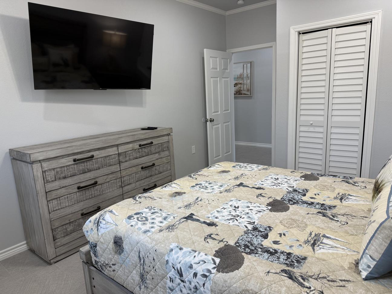 Coastal-inspired bedroom with a patterned quilt, mounted flat-screen TV, weathered wood dresser, white shutter closet doors, gray walls, and light tile flooring.
