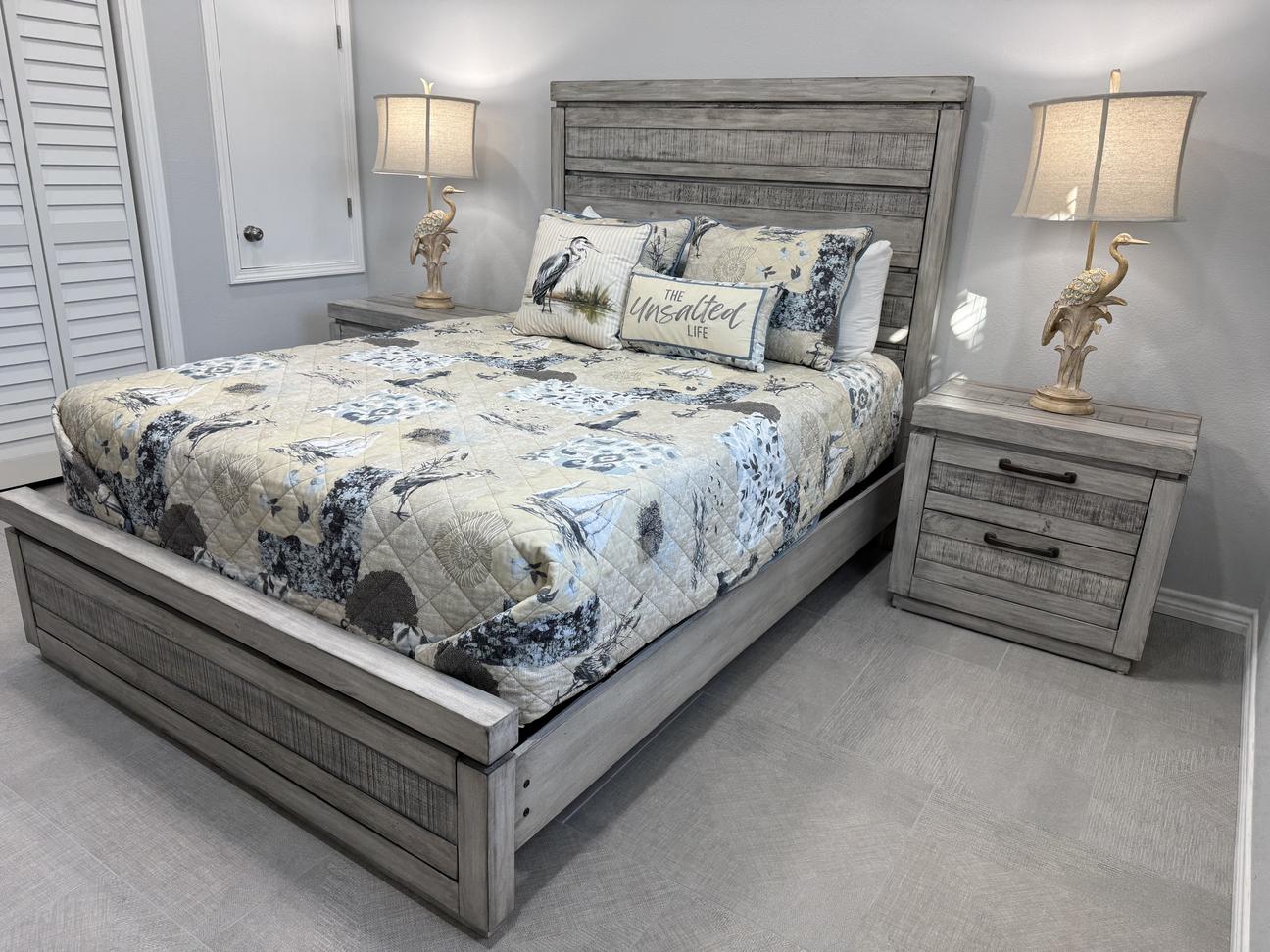Coastal-themed bedroom with a gray wood bed frame, matching nightstands, bird-themed lamps, patterned bedding, and light gray tile flooring with soft neutral walls.