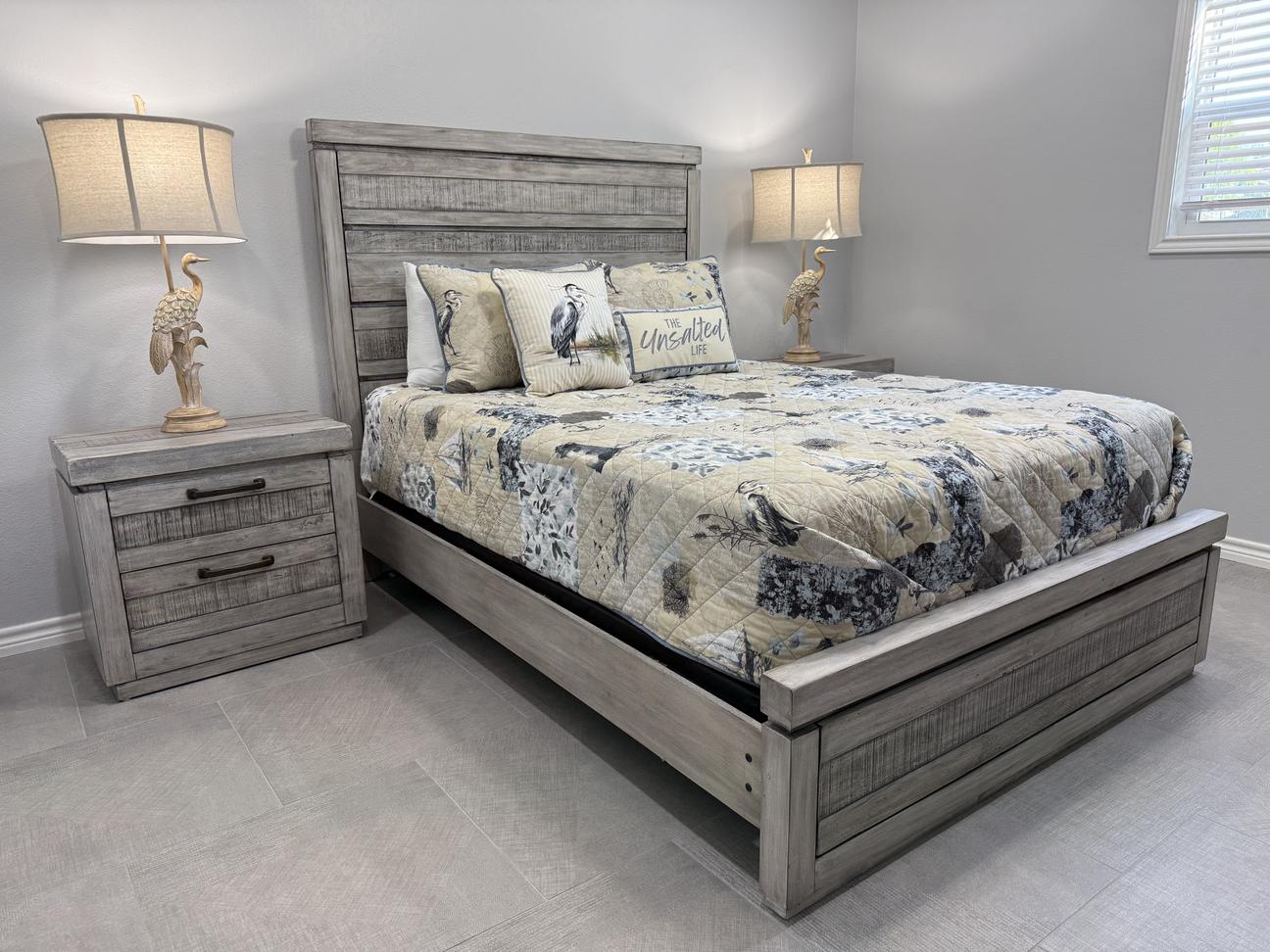Coastal-style bedroom with a gray wood bed frame, matching nightstand, bird-shaped lamps, patterned bedding with seaside decor, light gray walls, and tile flooring.