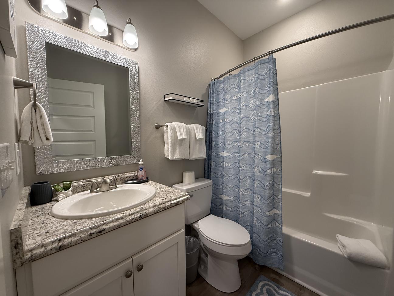 Modern coastal-style bathroom with granite vanity, decorative framed mirror, tub and shower combination, blue fish-pattern shower curtain, and soft neutral décor in a bright vacation rental.