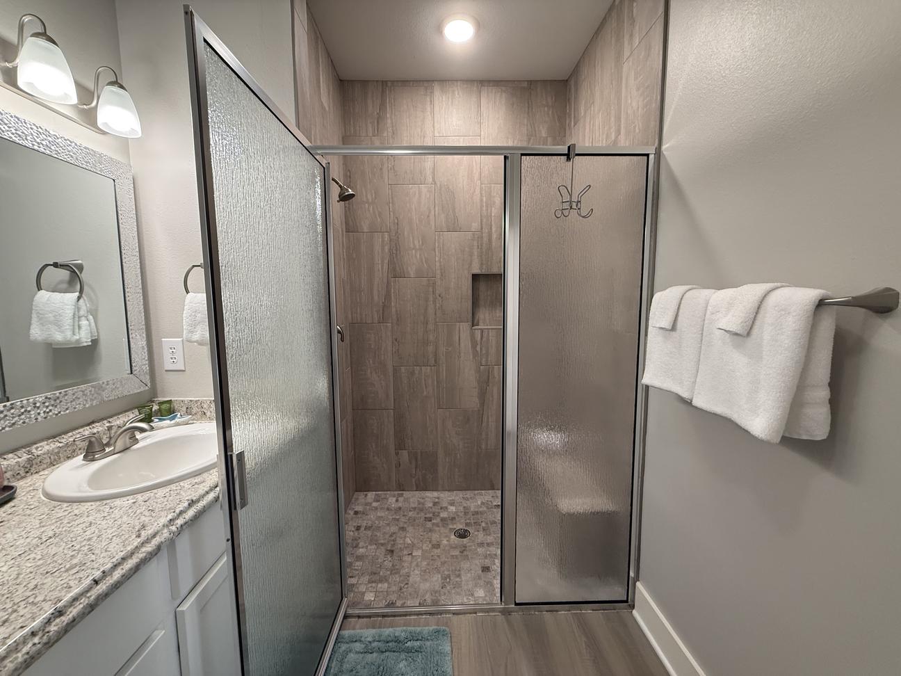 Modern coastal-style bathroom featuring a large tiled walk-in shower with glass doors, granite vanity countertop, fresh white towels, and soft neutral finishes in a vacation rental.