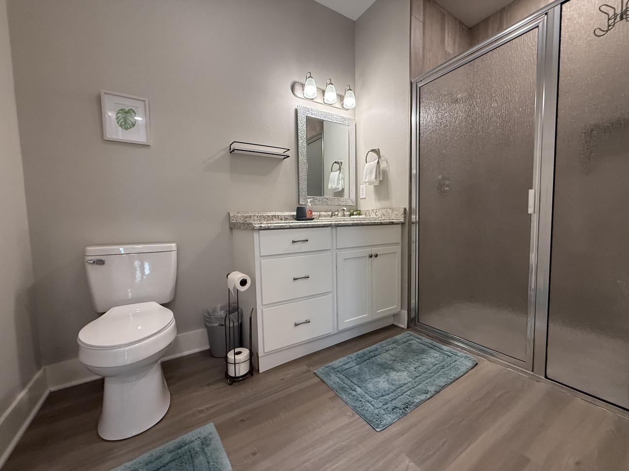 Modern coastal-style bathroom with large walk-in shower, white vanity with granite countertop, soft neutral décor, and light wood-look flooring in a bright vacation rental.
