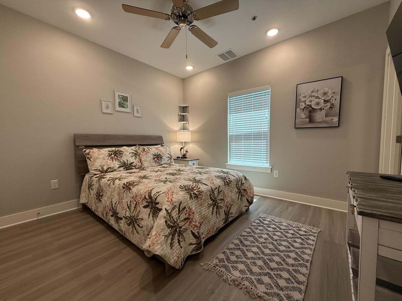 Spacious coastal-style bedroom with king bed, tropical-pattern bedding, ceiling fan, soft neutral walls, bedside lighting, and wood-look flooring in a modern vacation rental.