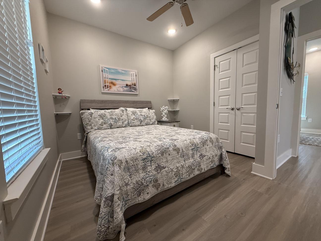 Coastal-inspired bedroom with queen bed, neutral beach-themed bedding, ceiling fan, wood-look flooring, and soft natural light in a modern vacation rental.