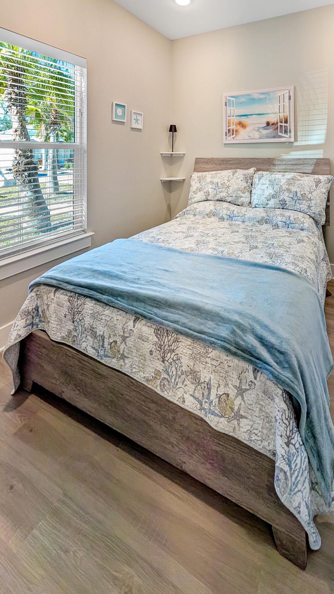 Queen bed with tan and blue coastal bedding next to a window with white shades. 