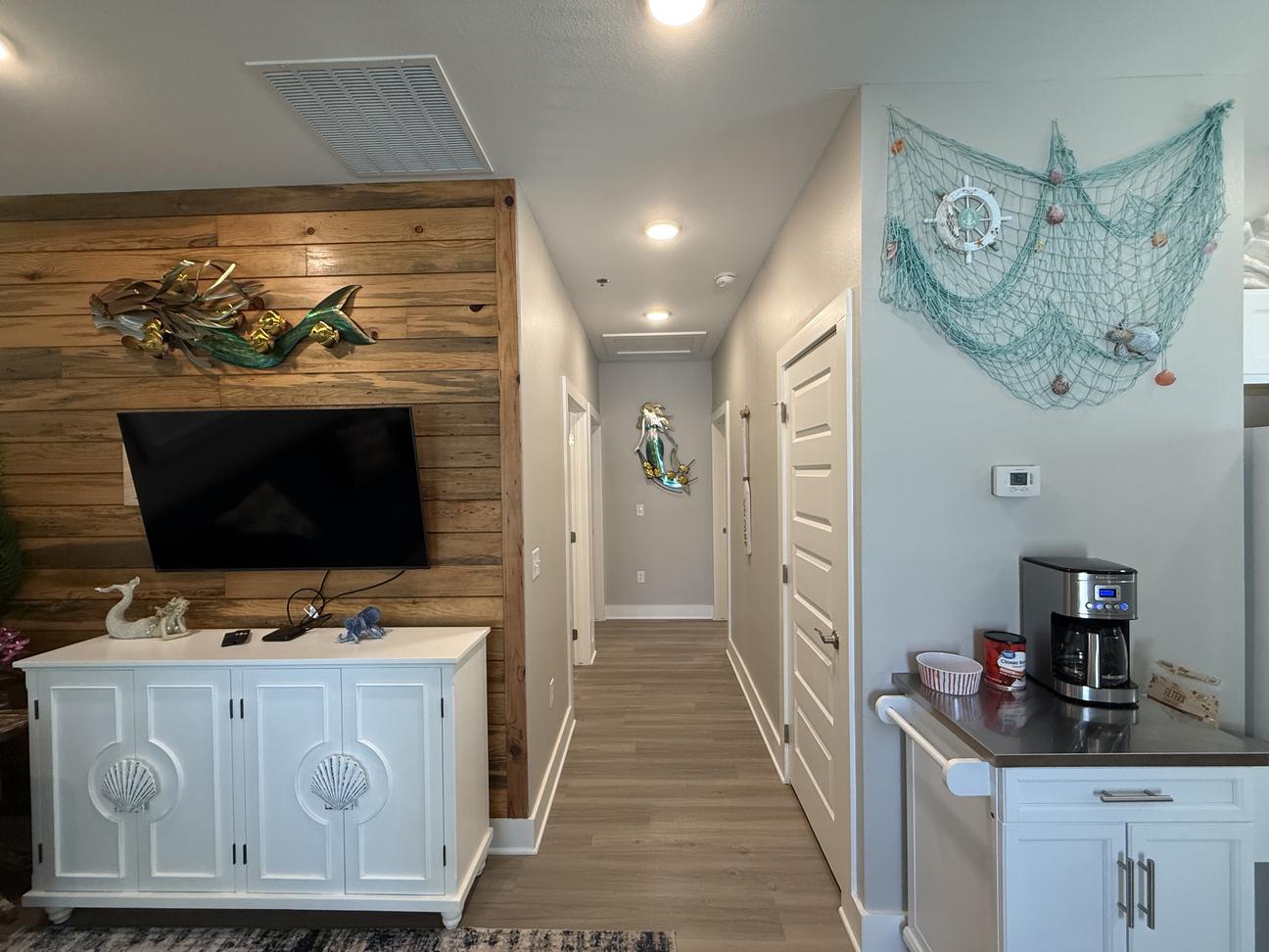 Coastal-inspired vacation rental interior with wood accent TV wall, beach-themed décor, coffee station, hallway leading to bedrooms, and light modern finishes throughout the space.