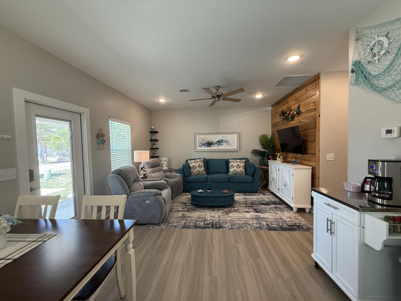 Open coastal-style living and dining area with comfortable seating, wood accent TV wall, dining table, coffee station, beach-inspired décor, and bright natural light throughout the vacation rental.