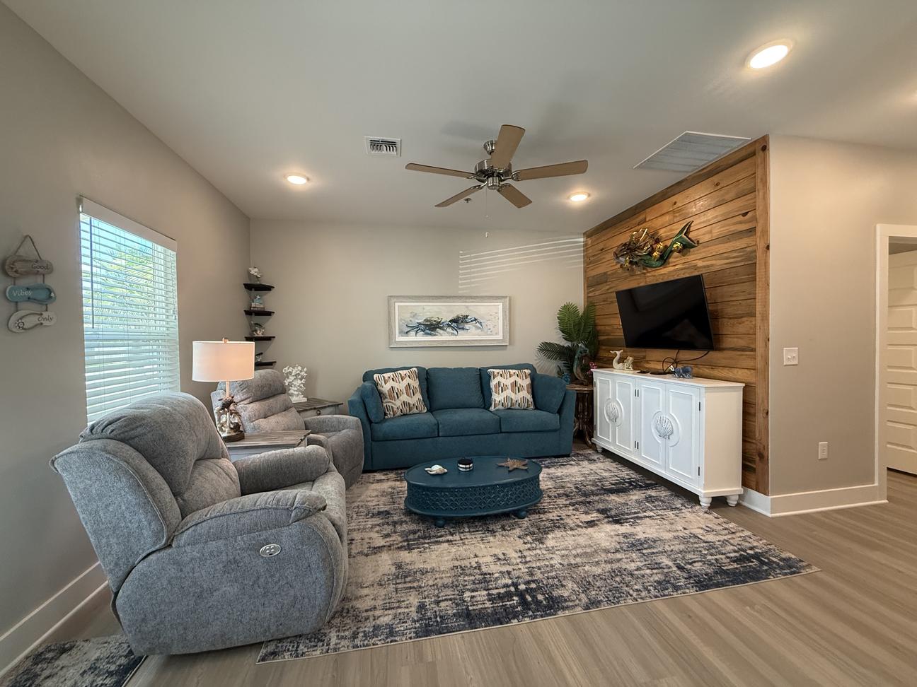 Coastal-inspired living room with blue sofa, gray recliners, wood accent wall, large flat-screen TV, ceiling fan, and beach-themed décor in a bright open-concept vacation rental.