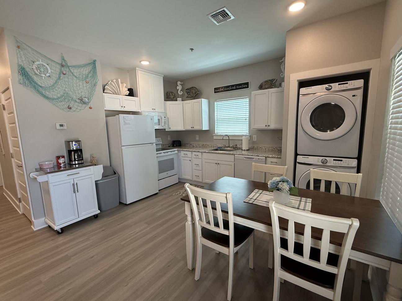 Open coastal-style kitchen and dining area with white cabinetry, full-size appliances, stacked washer and dryer, dining table for four, coffee station, and beach-inspired décor in a bright vacation rental.